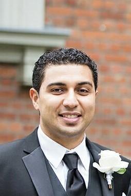 Sam Abidir smiles, wearing a formal suit with a white boutonniere. He is in front of a brick wall.